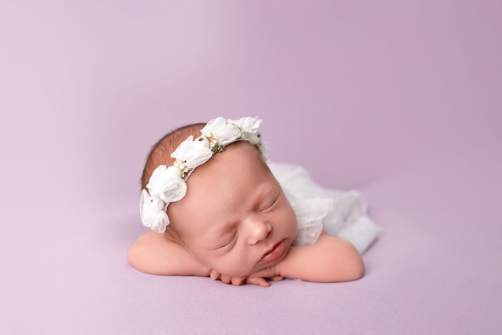 Peaceful newborn portrait with white floral crown against purple backdrop.