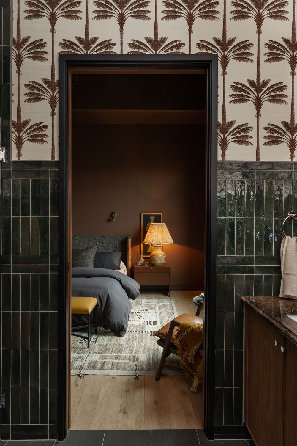 Cozy bedroom viewed through doorway with palm tree wallpaper, warm lamp, gray bedding, and green tile walls.