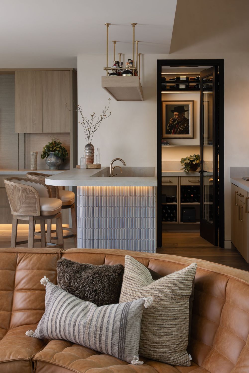 Modern open-plan kitchen with tan leather sofa, blue tile island, wine cellar, brass pendant light, and rattan bar stools.