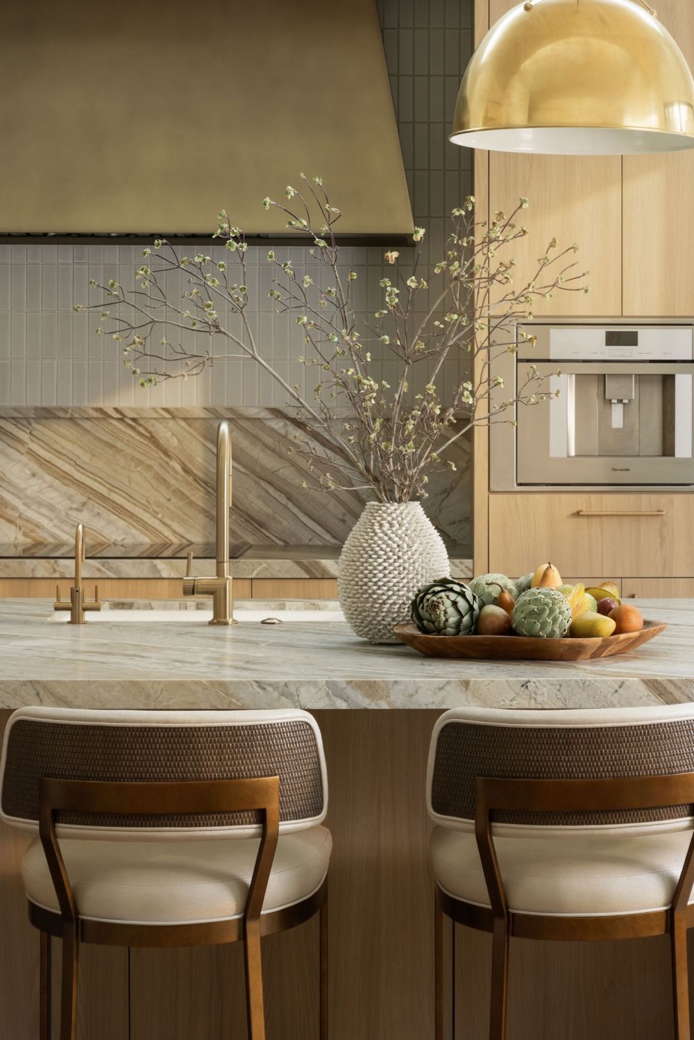 Elegant kitchen island with marble countertop, bar stools, vase with branches, fruit bowl, and warm wood cabinetry.