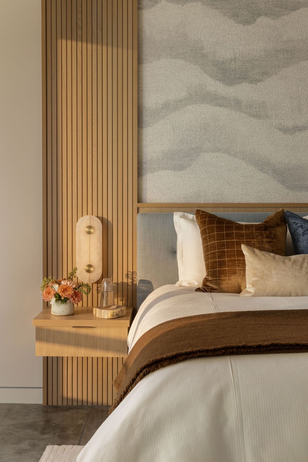 Modern bedroom with warm wood slat wall, floating nightstand, earth-tone pillows, and abstract mural headboard.