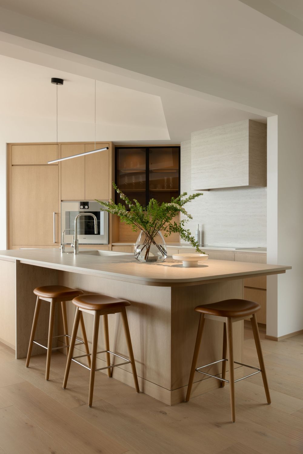 Modern minimalist kitchen with wooden island, bar stools, pendant light, built-in oven, and fresh fern centerpiece.