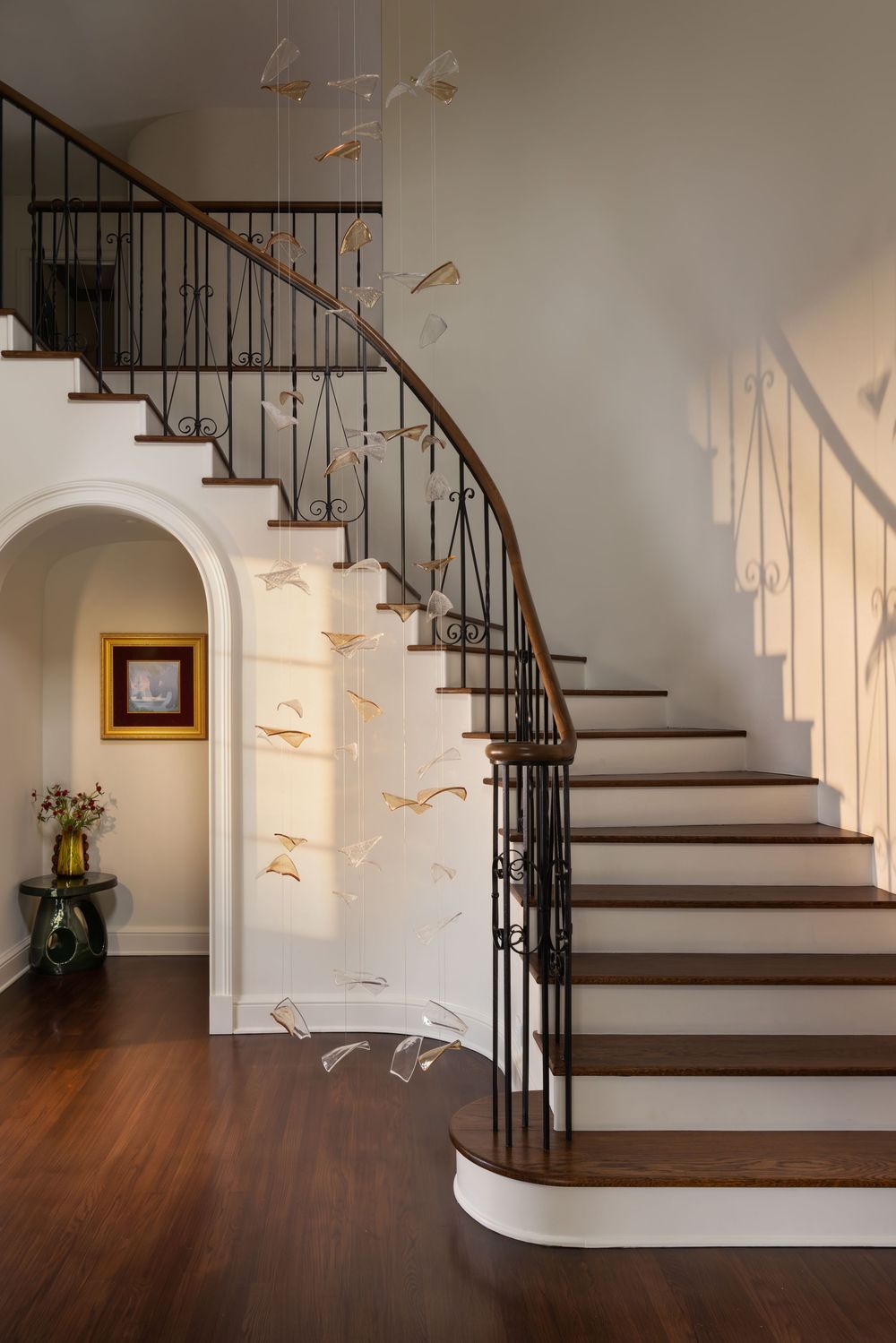 Elegant curved staircase with wrought iron balusters, wood handrail, and bird sculpture chandelier in a bright foyer.