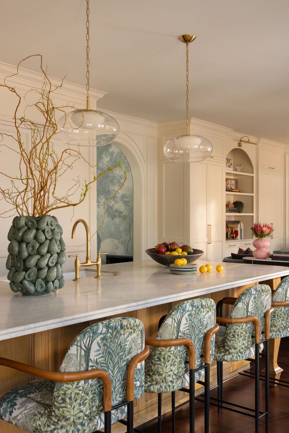 Elegant kitchen with marble island, green floral barstools, brass pendant lights, and a textured sage vase with branches.