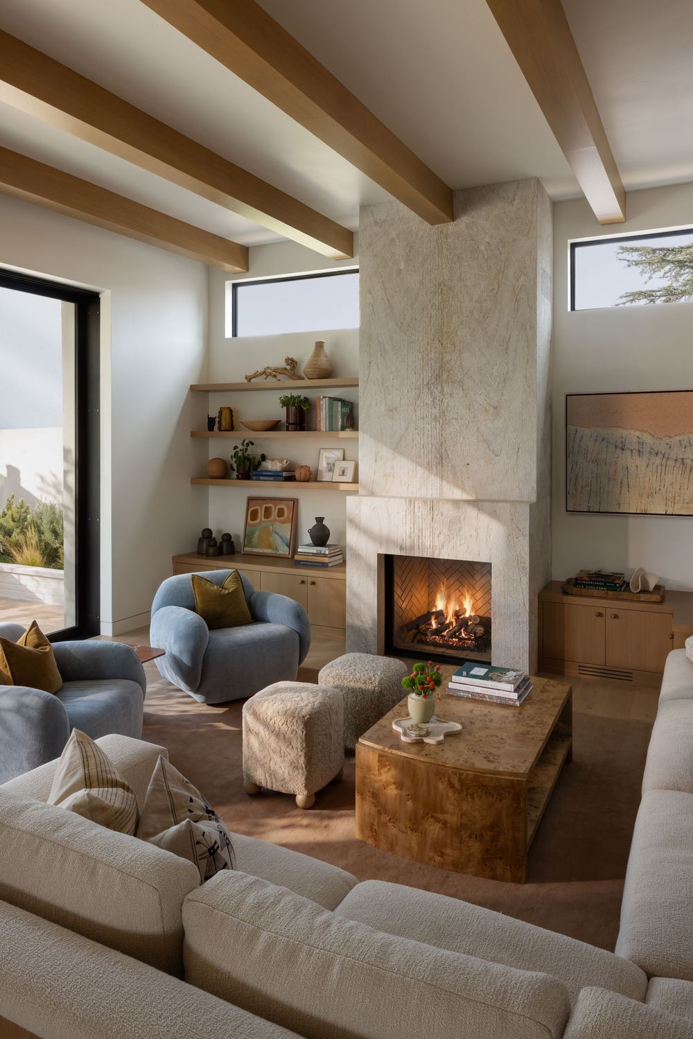 Cozy living room with a travertine fireplace, exposed wood beams, blue armchair, burl wood coffee table, and neutral sofa.