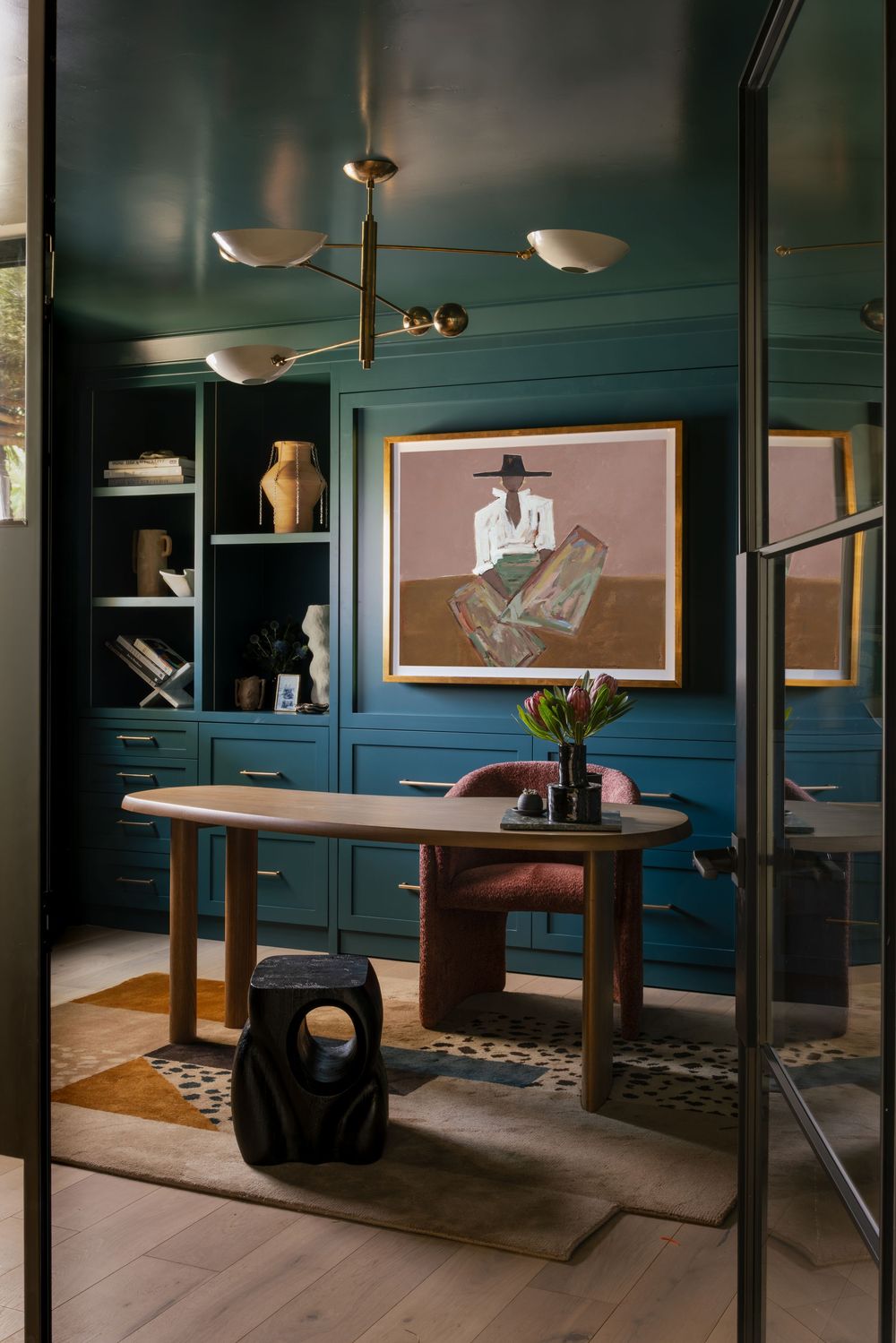 Elegant teal home office with built-in cabinets, oval wood desk, brass chandelier, and framed figurative artwork.