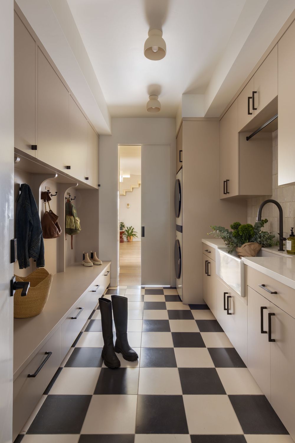 Modern laundry mudroom with black-and-white checkered tile floor, cream cabinetry, stacked washer-dryer, and black knee-high boots.