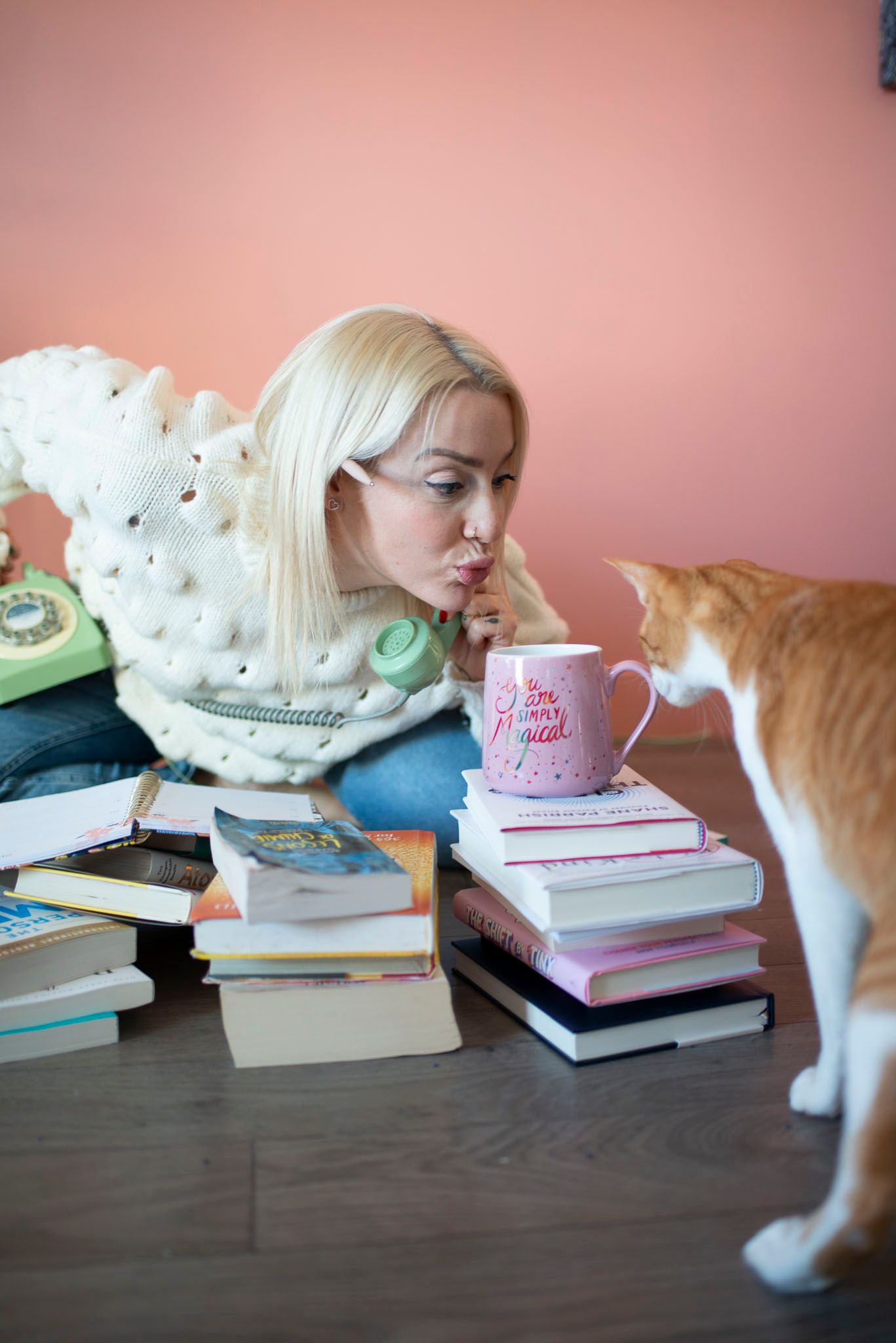 Woman in white sweater studying with books while cat investigates her pink coffee mug against coral wall.