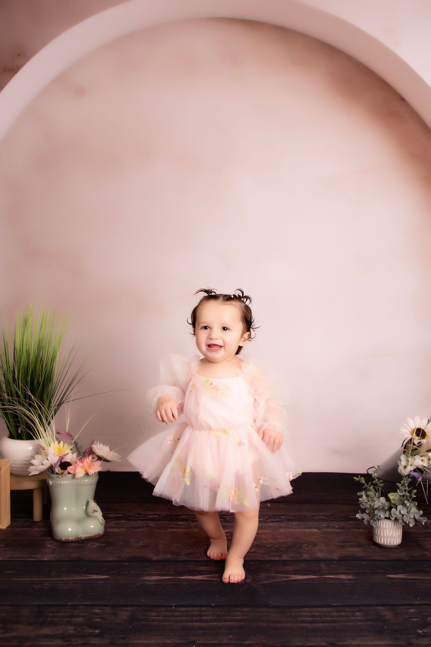 A toddler in a pink tulle dress dances in a minimalist studio setting with potted plants and flowers.