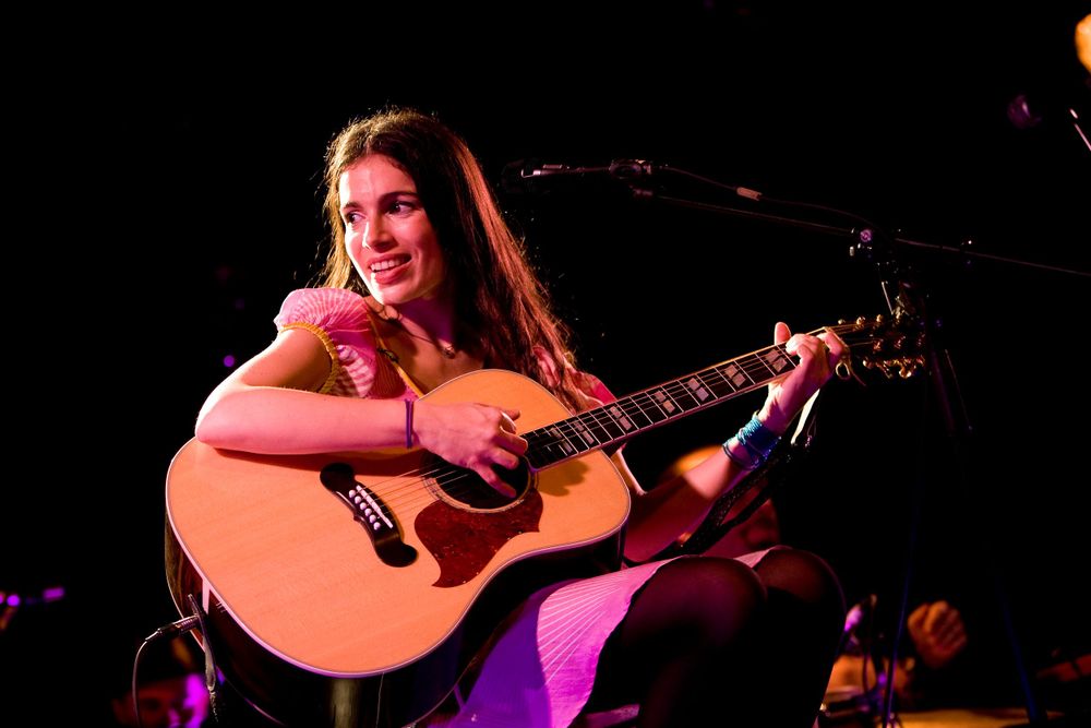 Musician playing acoustic guitar on stage under warm spotlights during a live performance.