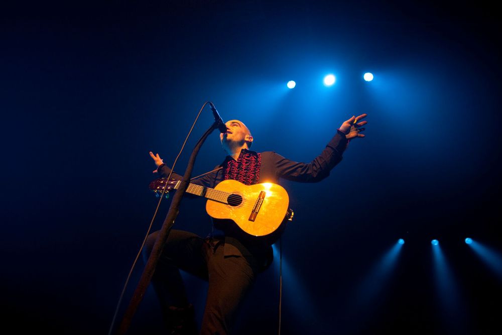 Performer with acoustic guitar silhouetted against dramatic blue stage lighting.