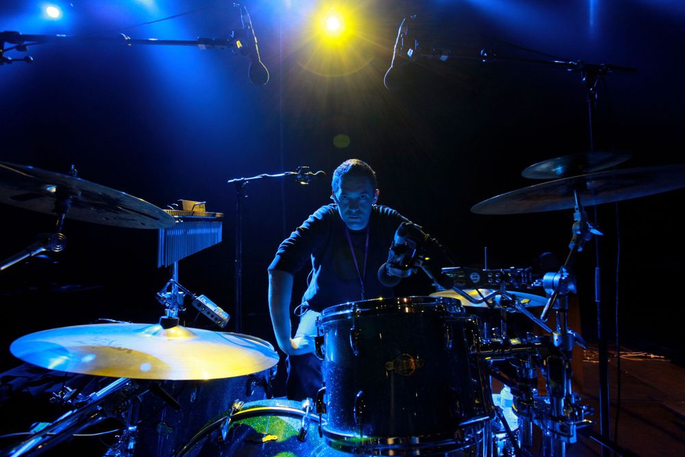 Drummer performs on stage with dramatic blue and yellow concert lighting.
