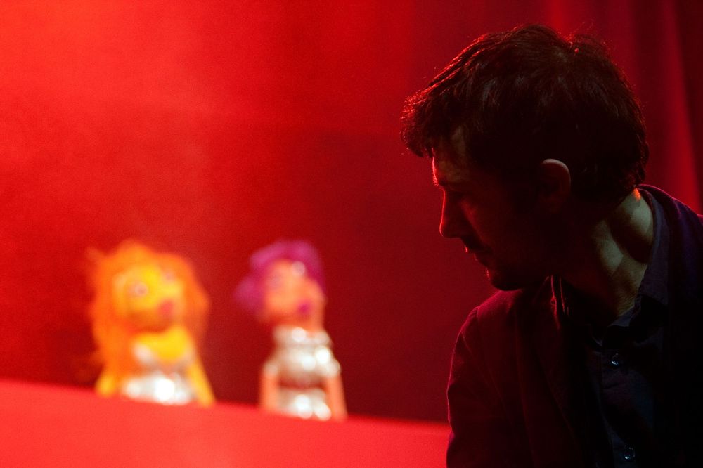 Silhouette of puppeteer performing with colorful puppets against red backdrop.