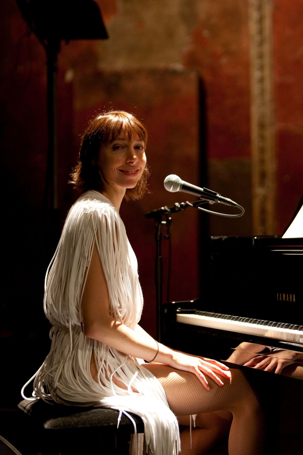 Performer in white dress seated at piano with microphone in warm-lit venue setting.