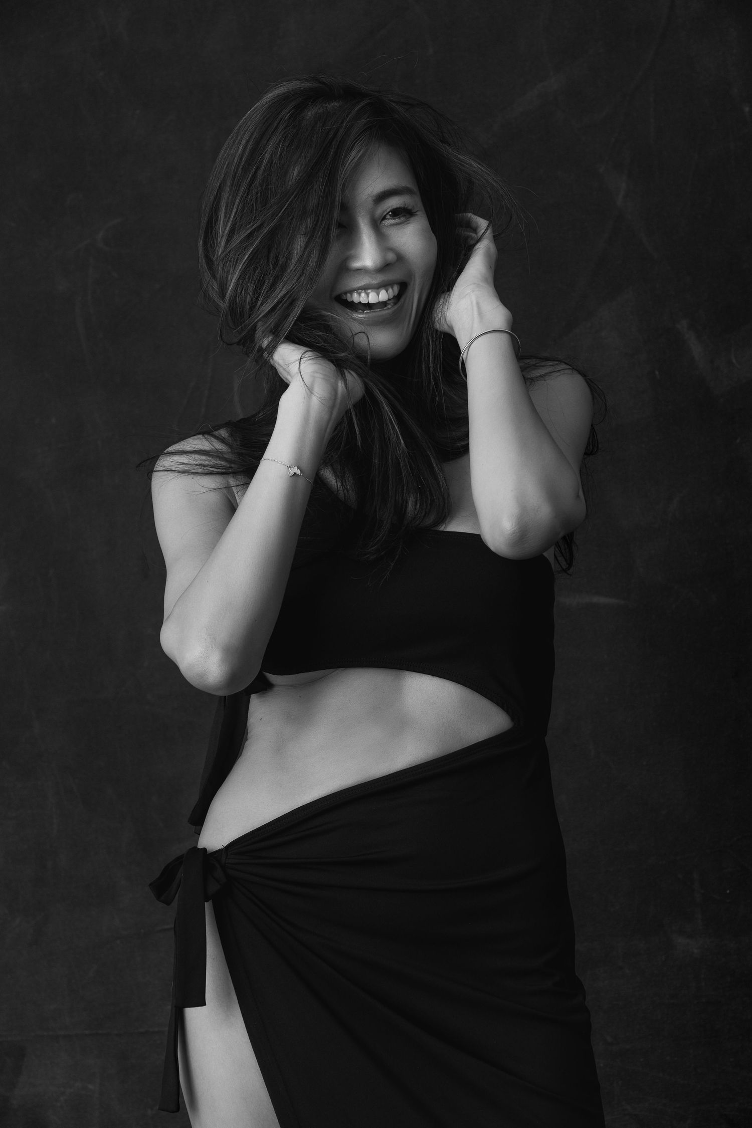 Black and white portrait of a joyful woman in dark crop top and wrap skirt posing against dark background.
