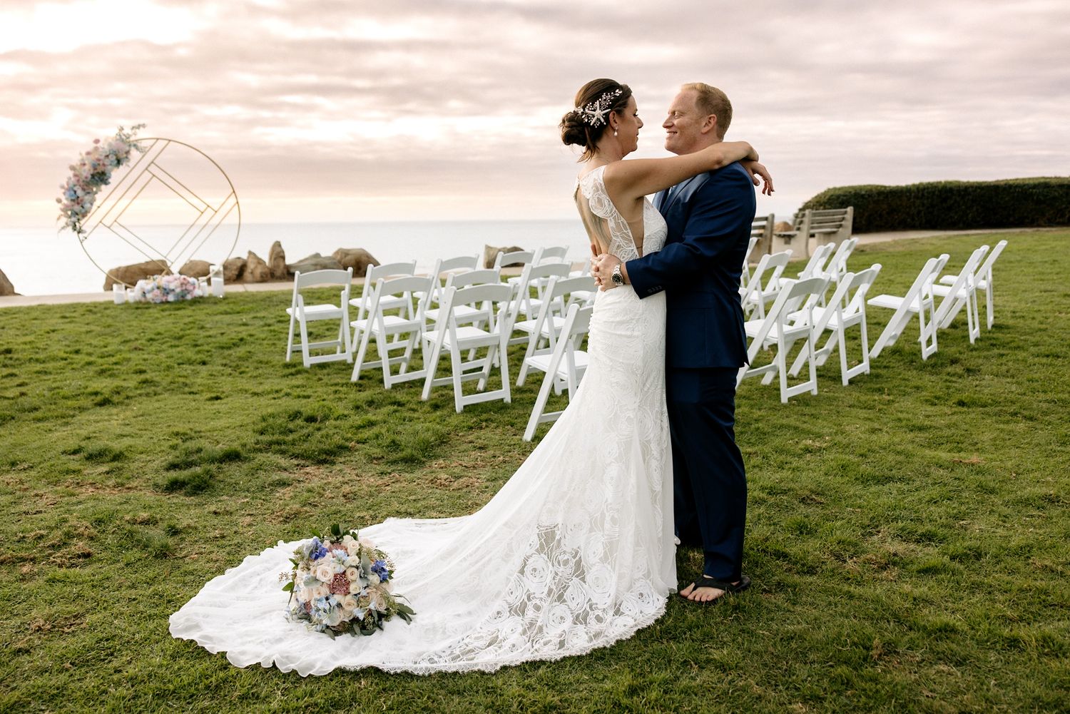 A couple embraces on a grassy hilltop with white chairs set up for an outdoor wedding ceremony in San Diego California.