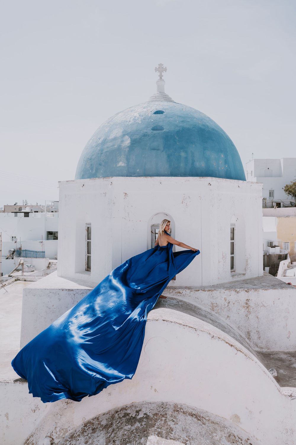 Dramatic blue dress flows in wind against white church with blue dome in Santorini.
