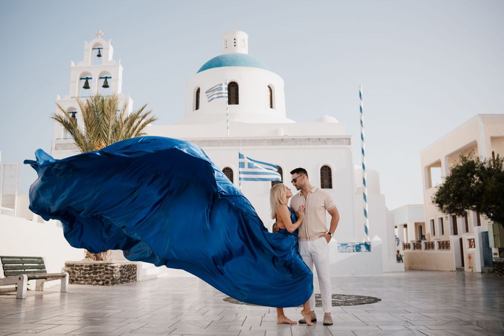 A flowing navy blue dress fabric catches wind in front of a classic white and blue Santorini church.