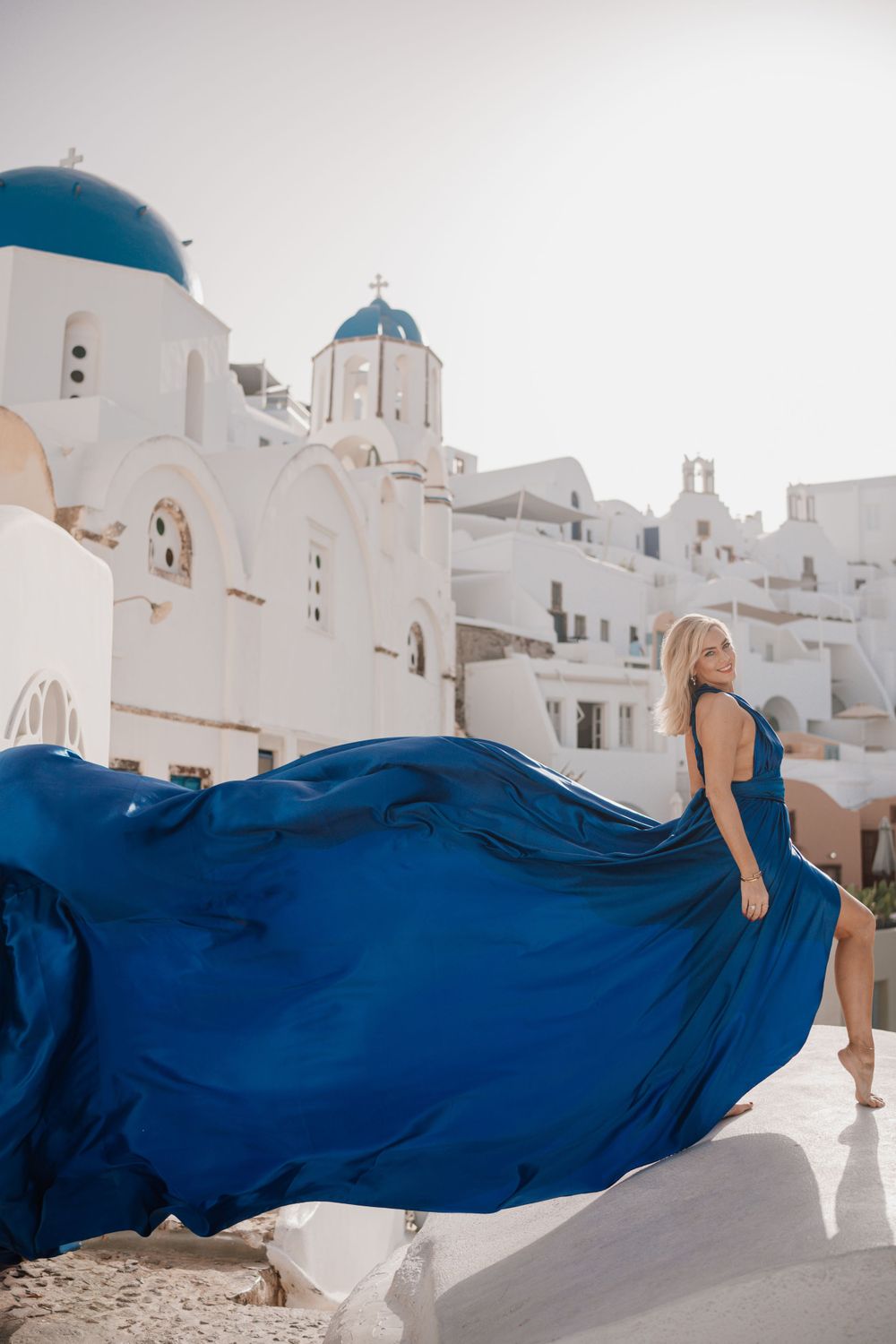 Elegant blue dress flowing dramatically against traditional white buildings with blue domes in Santorini.