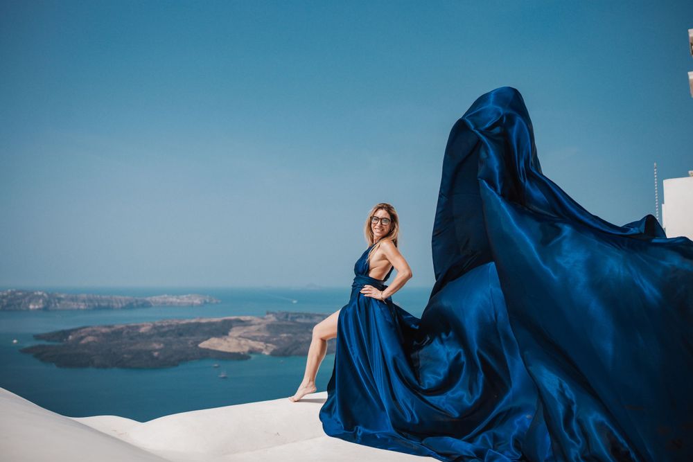 Long blue dress flowing in the wind against Santorini's stunning coastal backdrop.