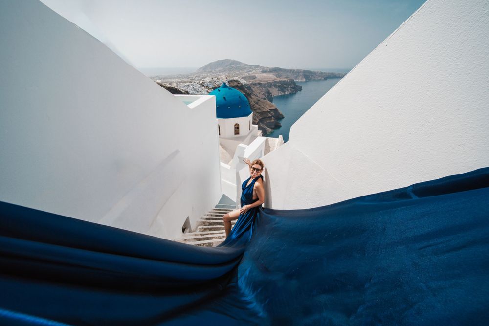 Blue flowing dress framed by white walls with classic Santorini architecture in background.