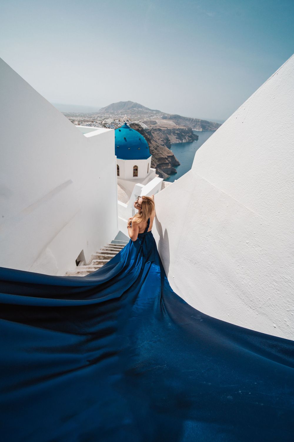 Dramatic blue dress cascading down white steps with iconic Santorini blue dome church view.