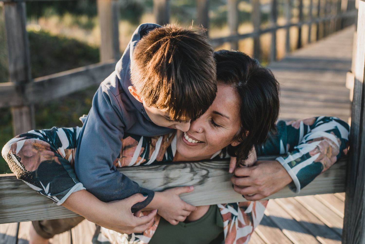 niño abraza a su madre desde atrás, ella está sentada en una pasarela de madera