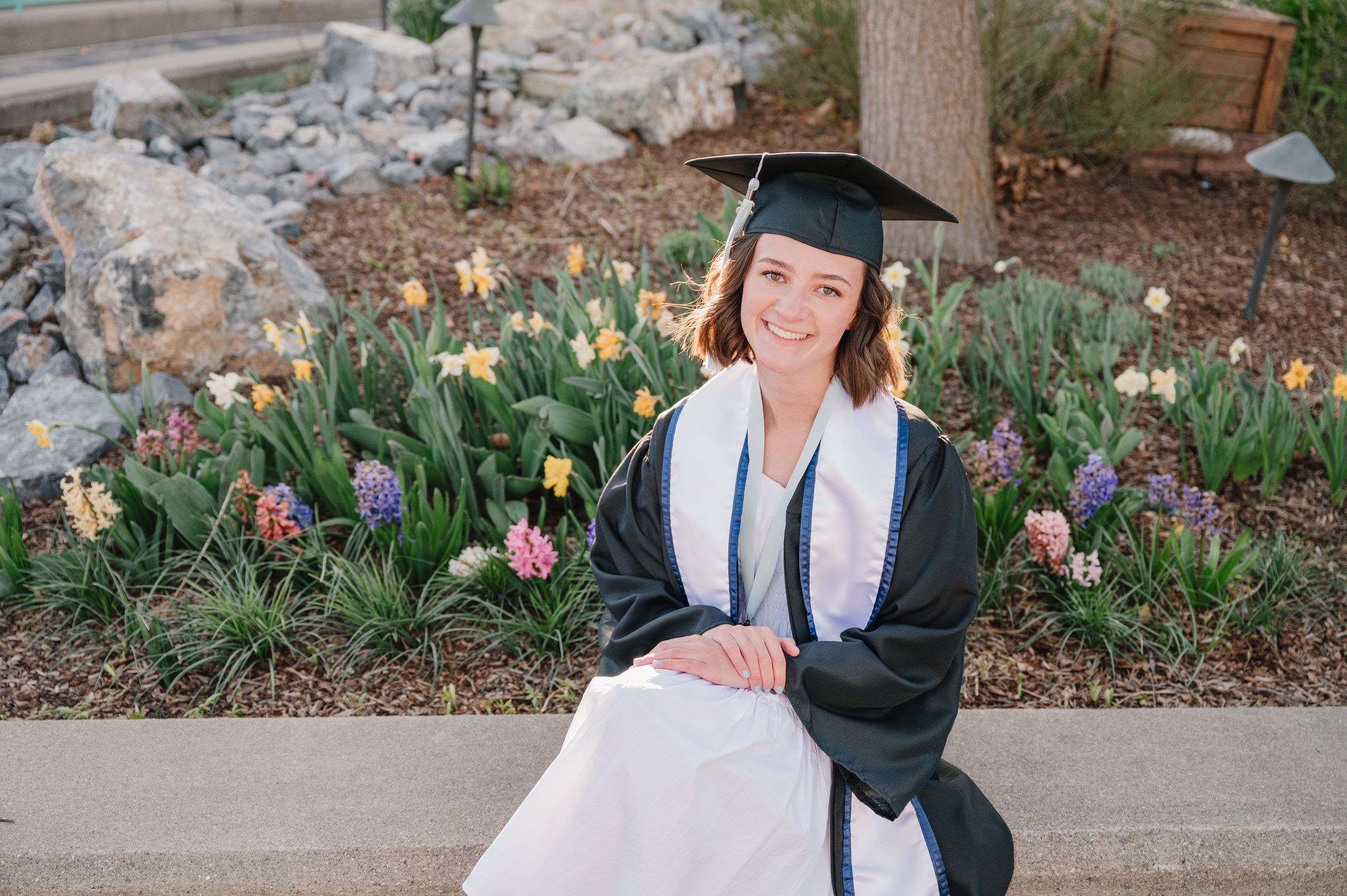 Graduate in cap and gown sitting on campus grounds with historic building in background.