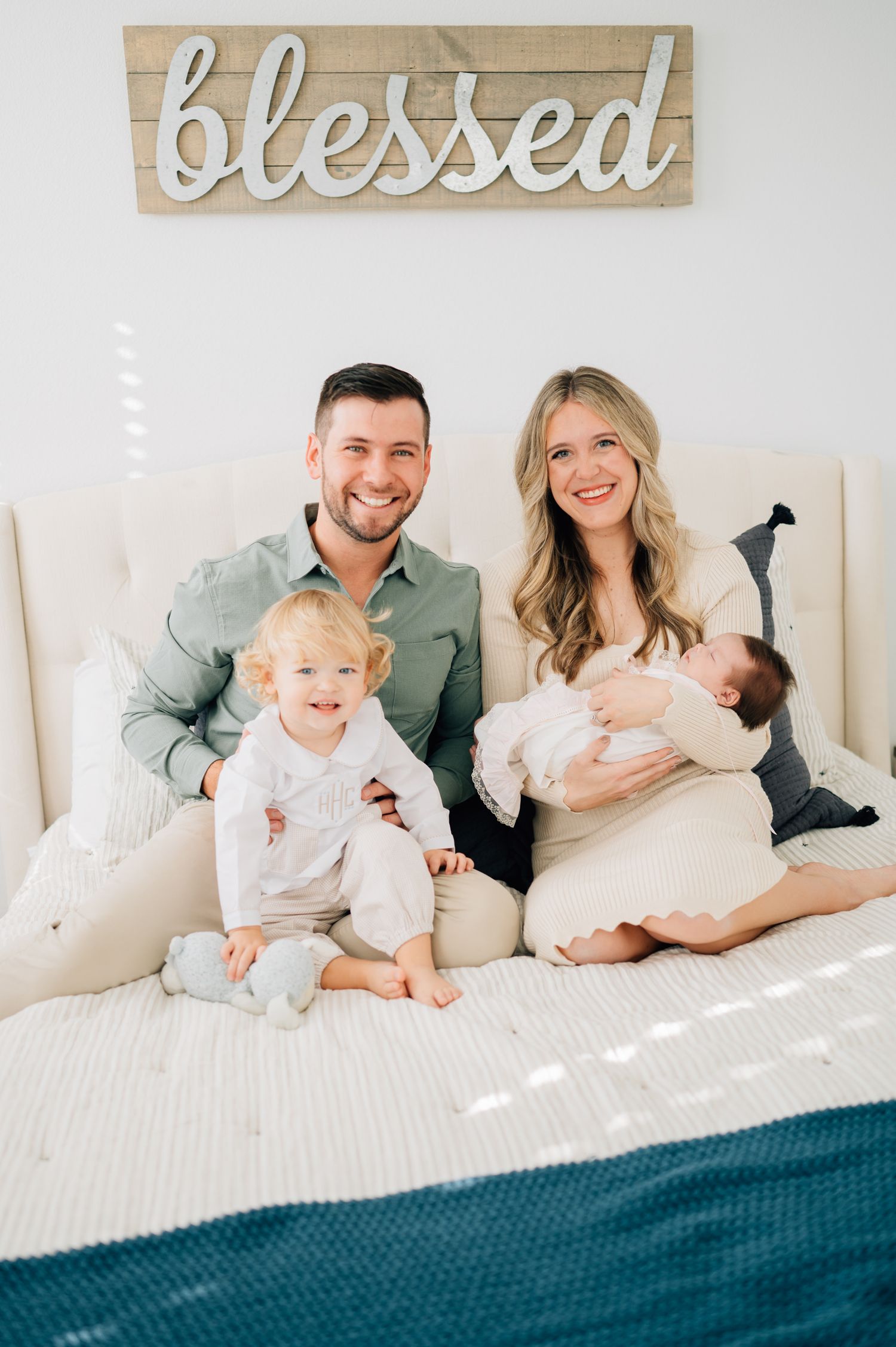 Family of four sitting on bed under wooden 'blessed' sign in bright room.