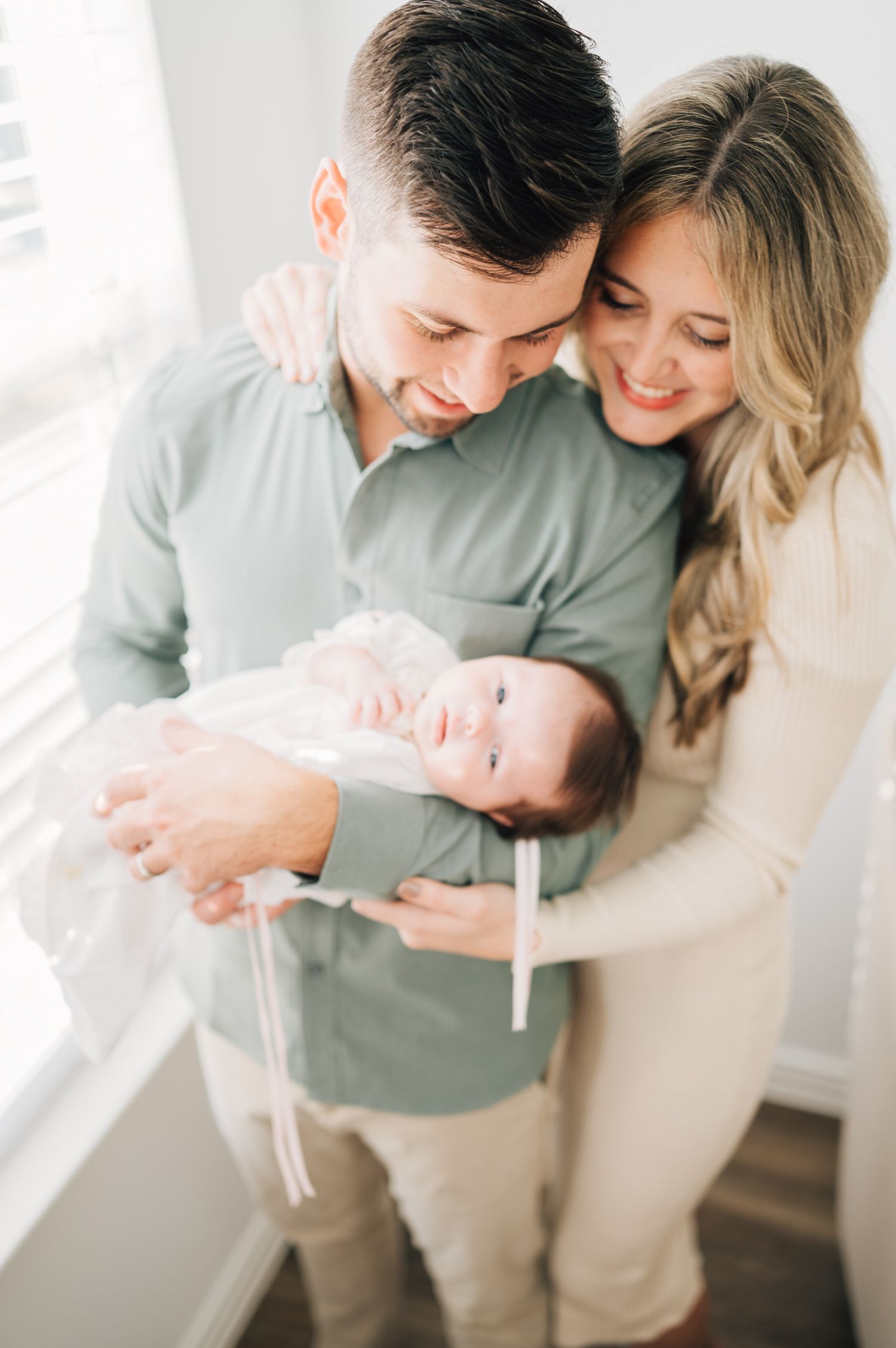 A couple in matching neutral tones embrace while holding their newborn baby in a light-filled room.