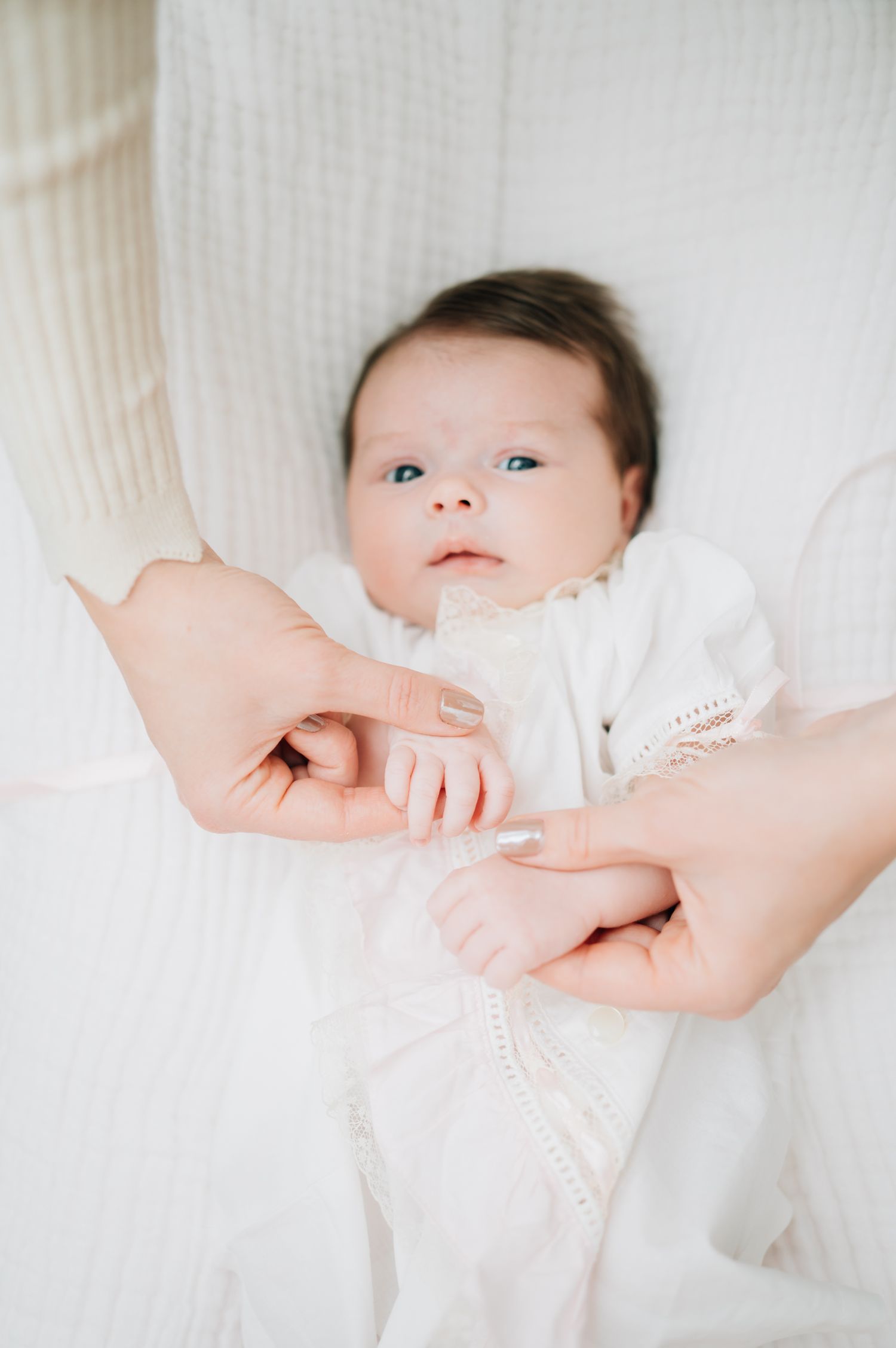 Close up of baby in white outfit reaching for parent's hand.