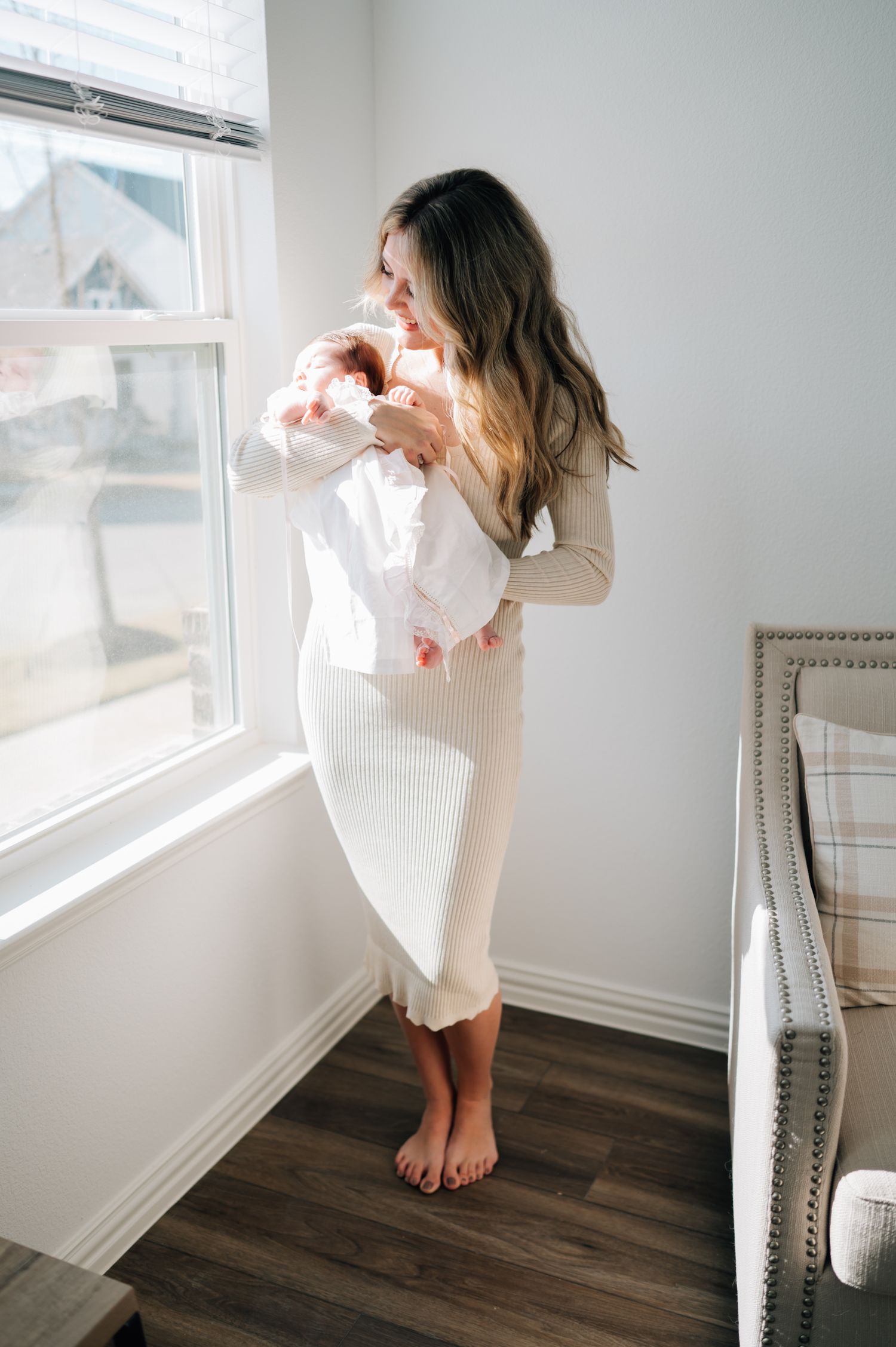 A woman in a cream dress holds her baby by a sunlit window in a modern room.