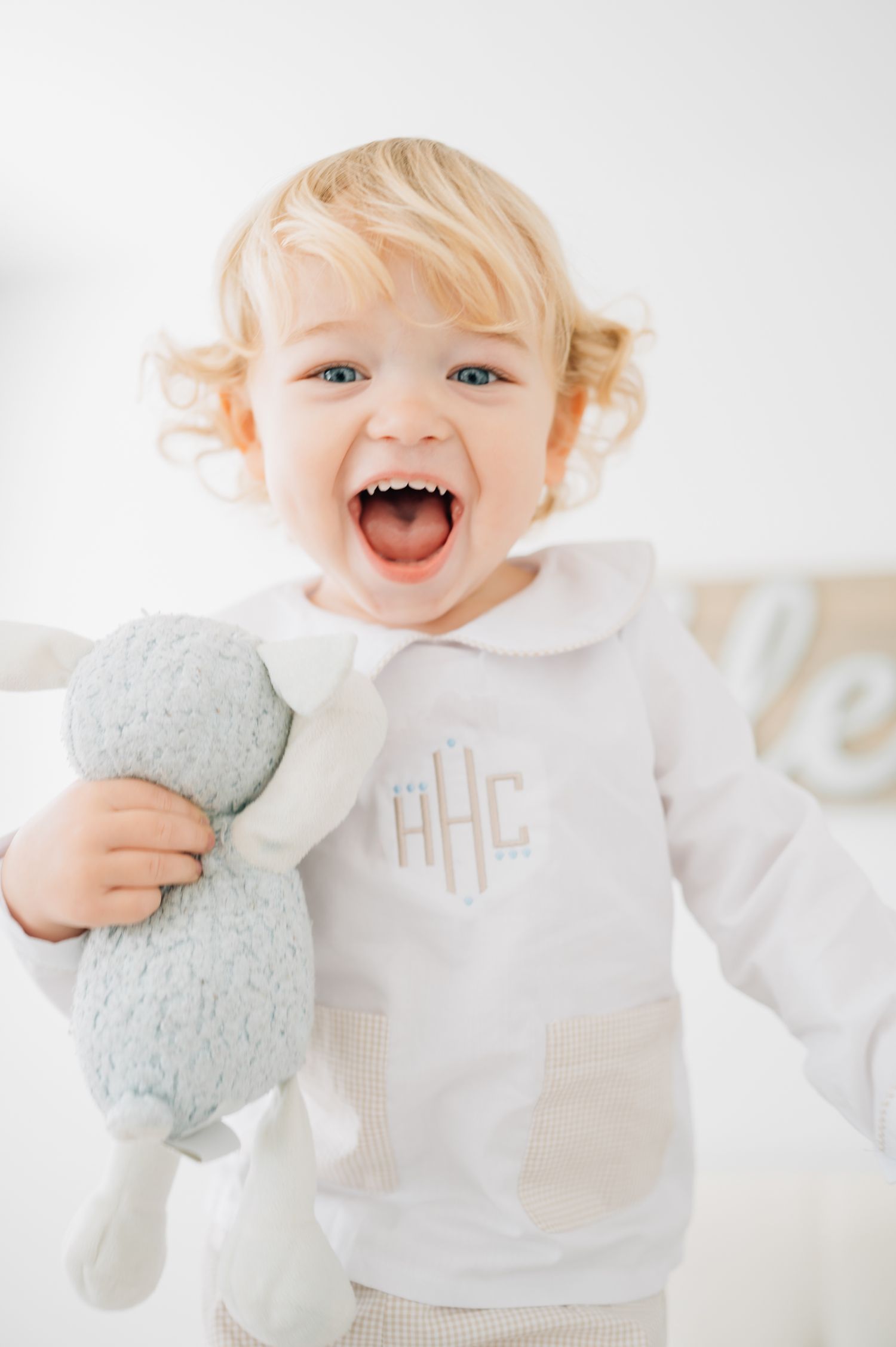 A joyful toddler in white holds a stuffed toy while laughing against a bright background.