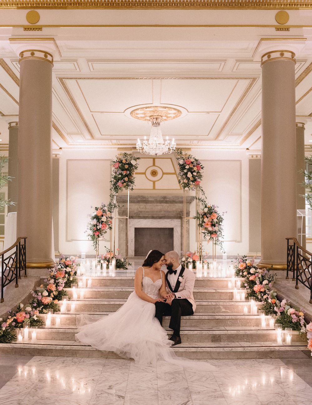 Bride and groom sitting on the grand marble staircase at Windsor Ballroom surrounded by luxurious floral arrangements.