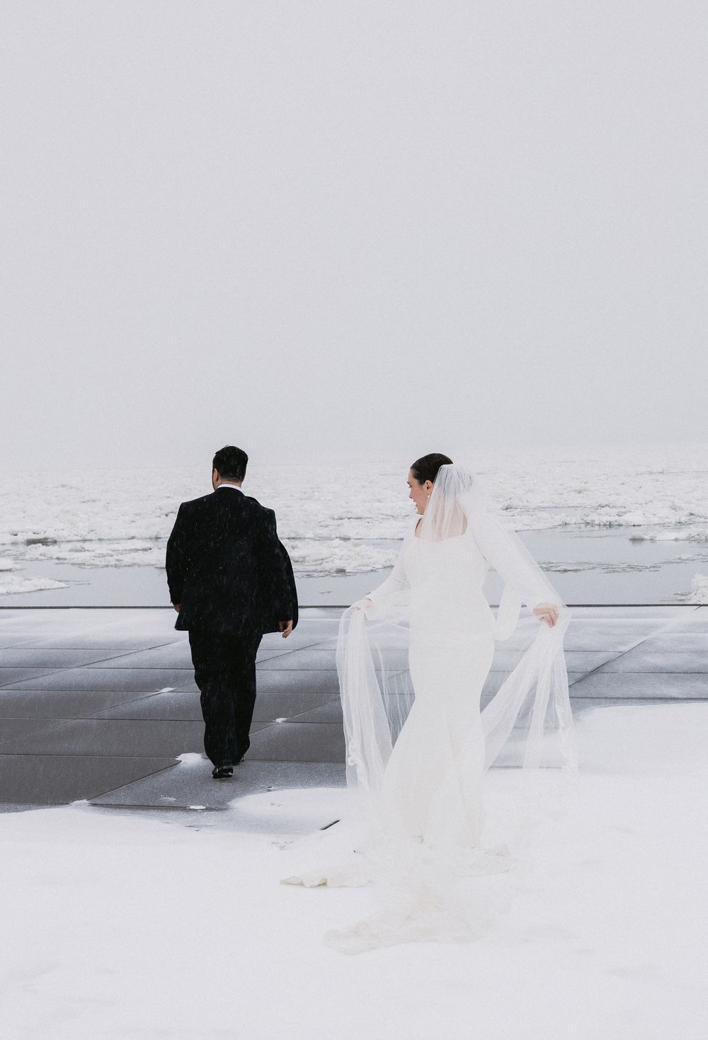 Bride in flowing white gown and veil walks with groom toward a frozen winter waterfront scene.