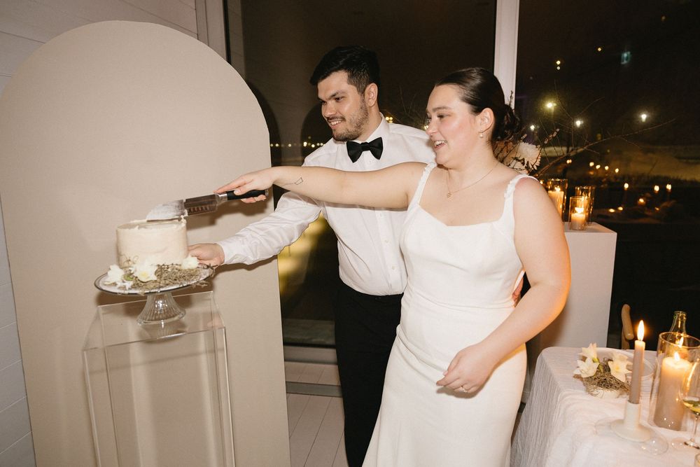 Smiling bride and groom in formal attire cut their wedding cake by a candlelit window at night.