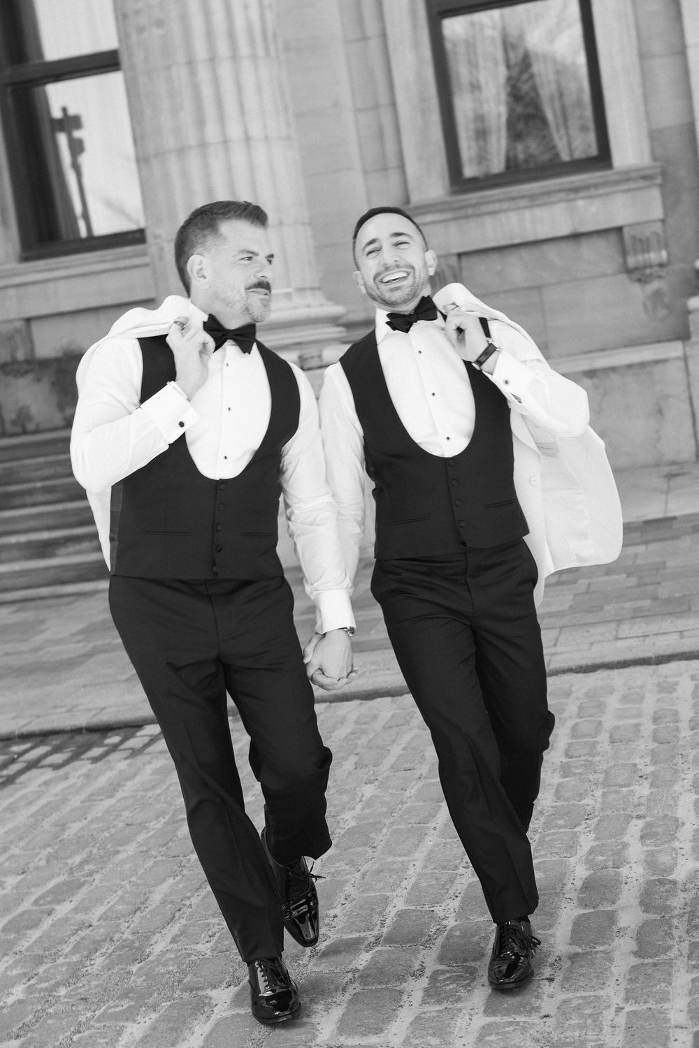 Two grooms in matching white tuxedo jackets and bow ties laughing while walking on cobblestone street.