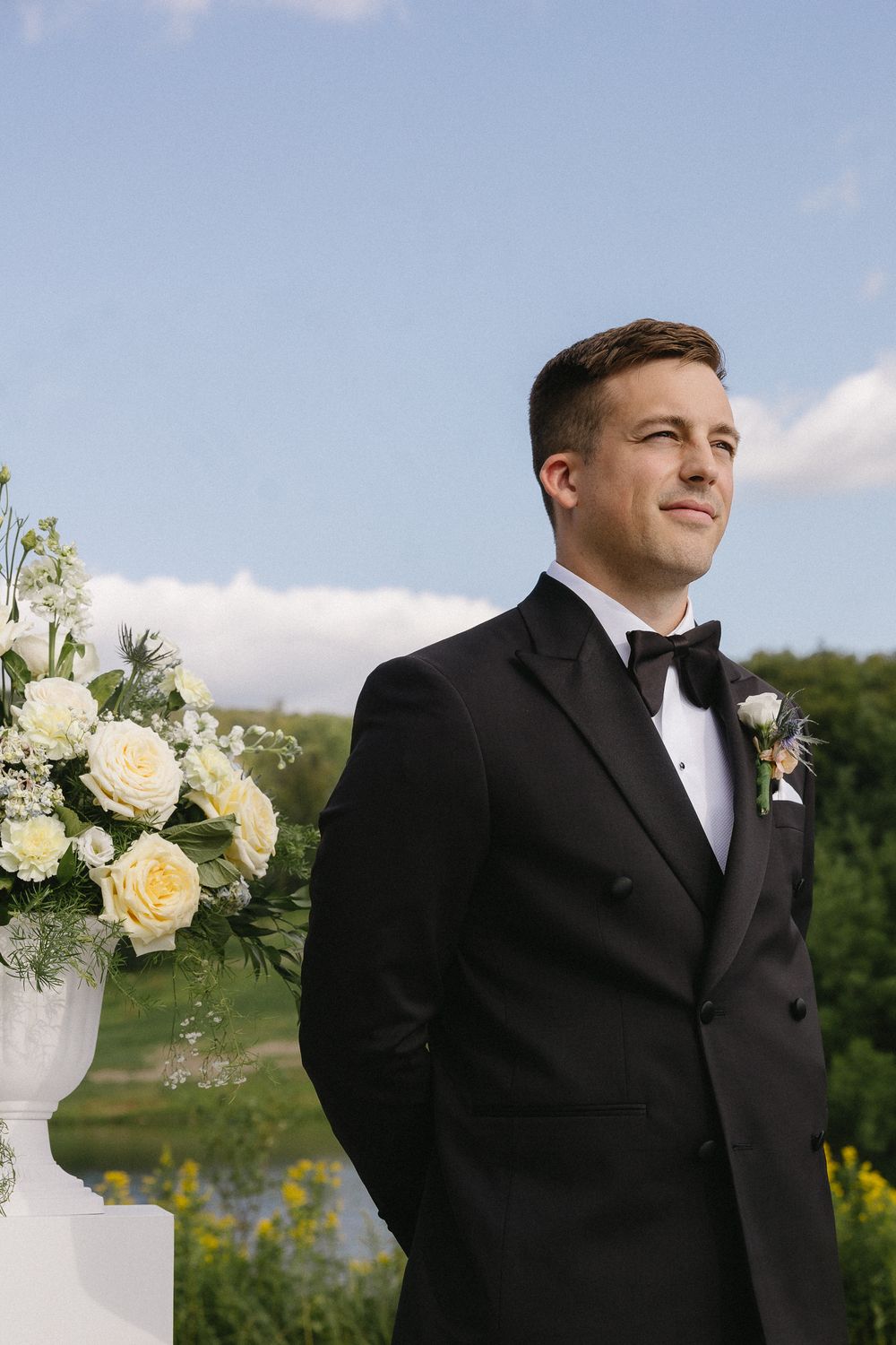 Groom in black tuxedo with bow tie and boutonniere standing at outdoor wedding altar with white roses.