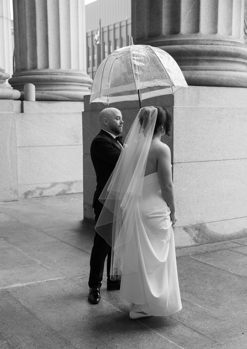 Black and white photo of bride and groom sheltering under clear umbrella beside stone column exterior.