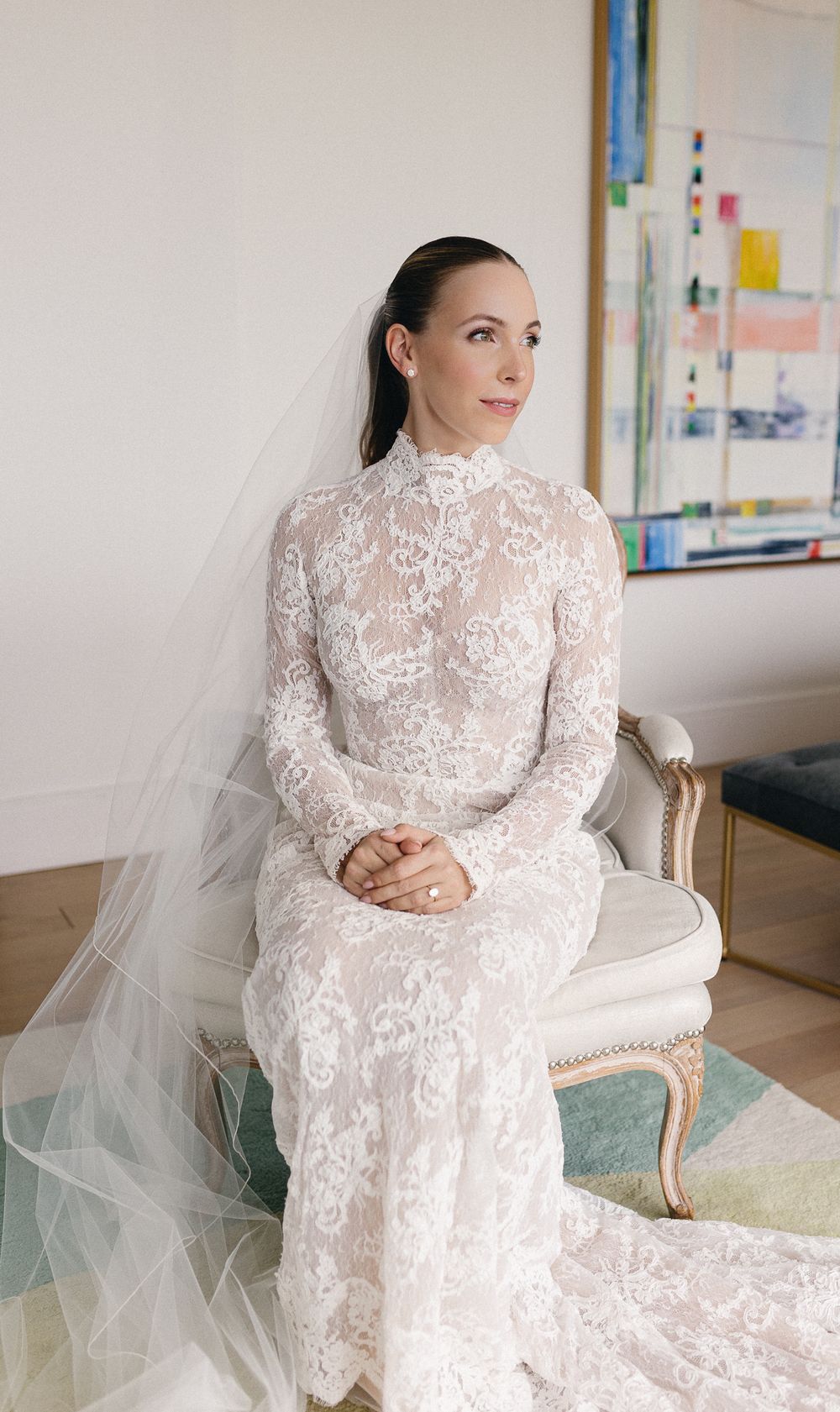 Bride in long-sleeve high-neck lace wedding gown with veil seated elegantly on white chair.