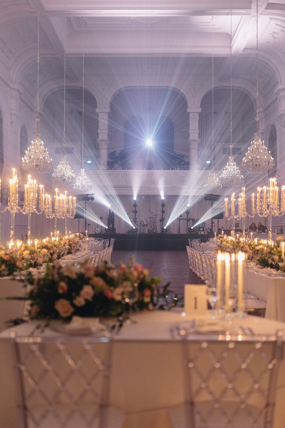 Grand ballroom wedding reception with crystal chandeliers, candlelit tables, and dramatic light beams.
