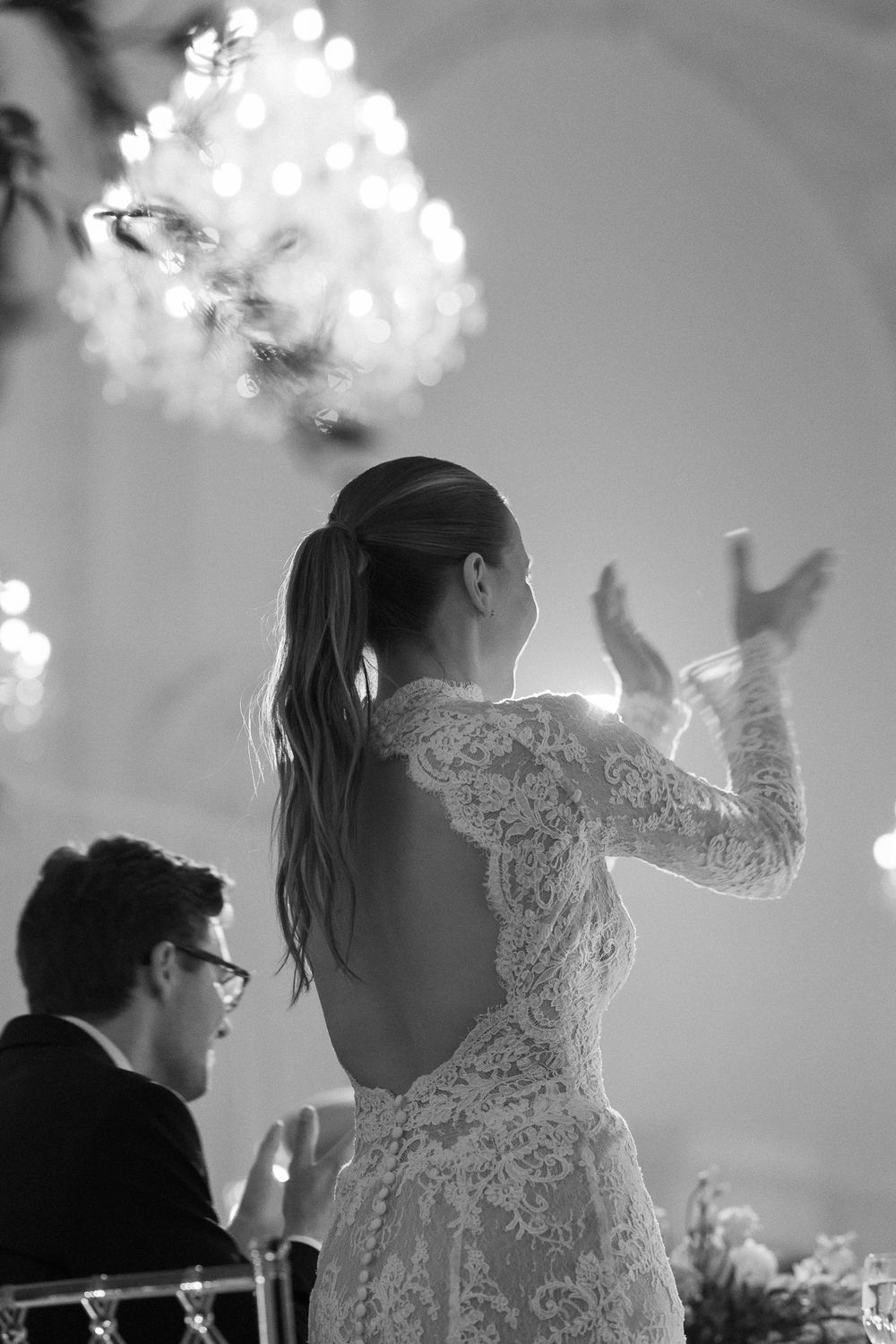 Bride in backless lace wedding gown gesturing during wedding reception with chandelier above.