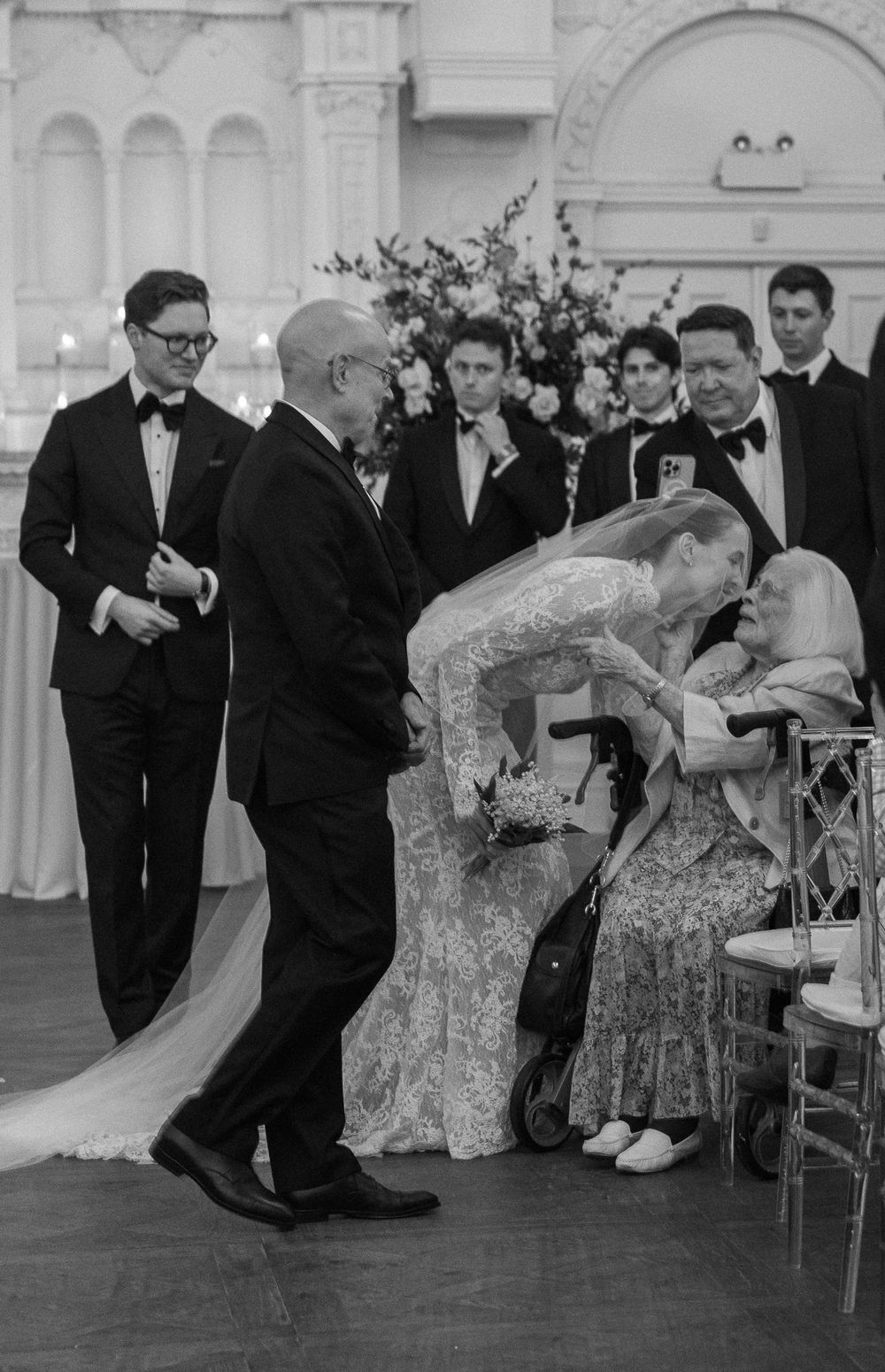 Black and white ceremony photo of bride in lace gown leaning to kiss elderly guest.