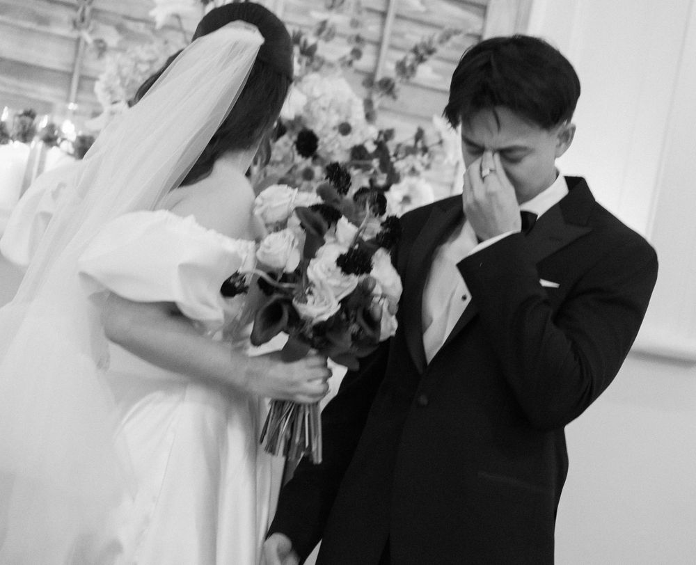 Groom wiping tears during emotional wedding ceremony while bride holds floral bouquet.
