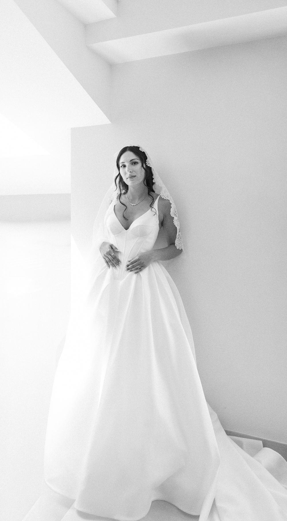 Elegant bride in a ballgown with lace veil poses in soft white light in a minimalist setting.
