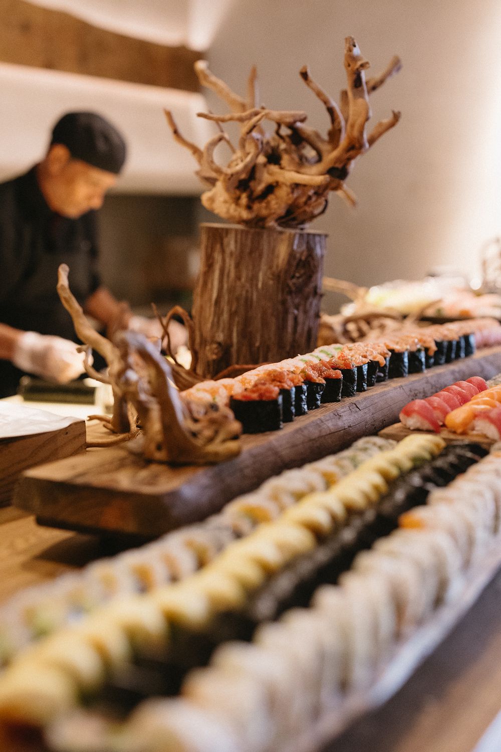 A sushi chef prepares an elaborate spread of assorted sushi rolls displayed on a wooden counter with decorative driftwood.