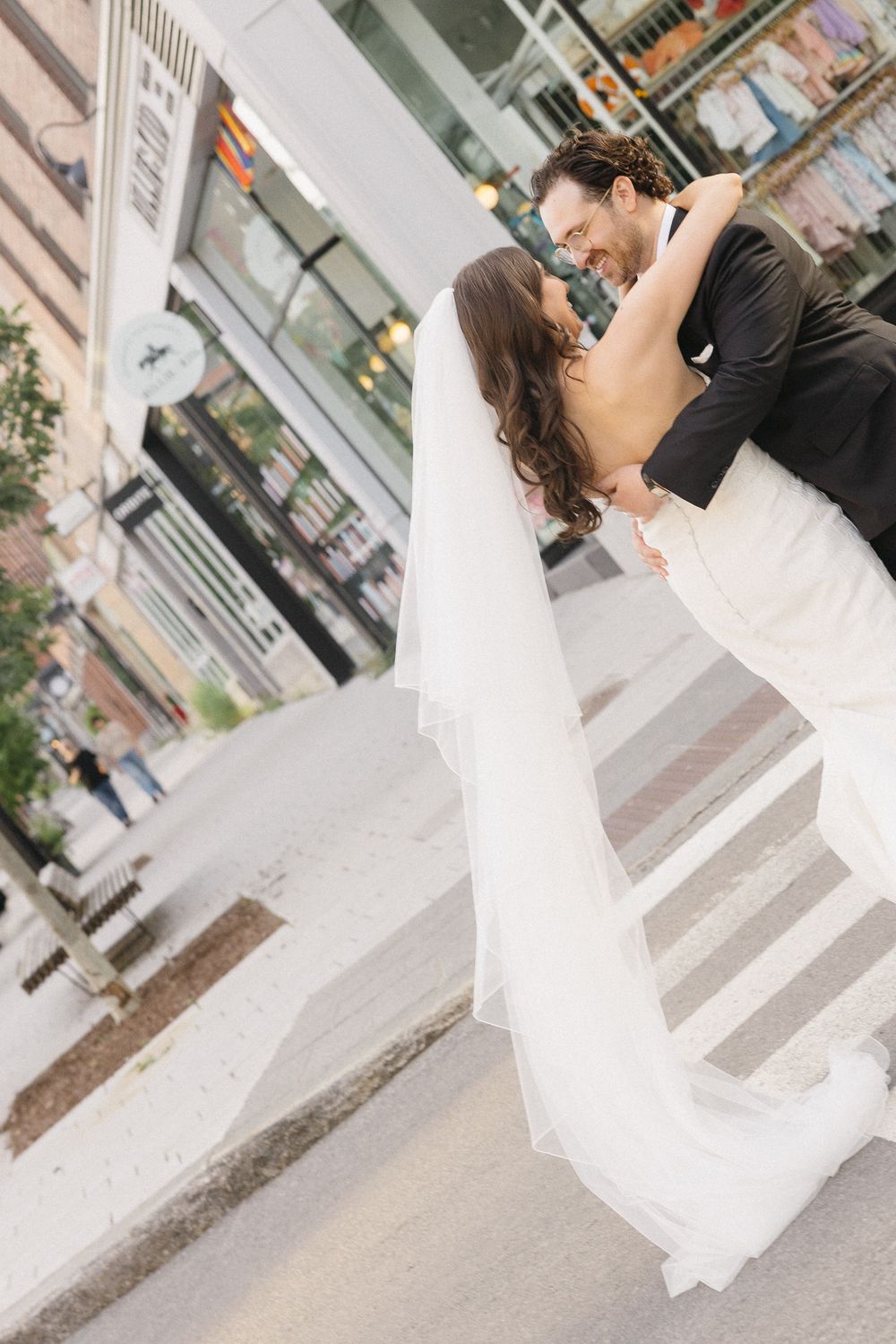Bride and groom share a romantic dip kiss on a city street, bride wearing a flowing white gown and veil.