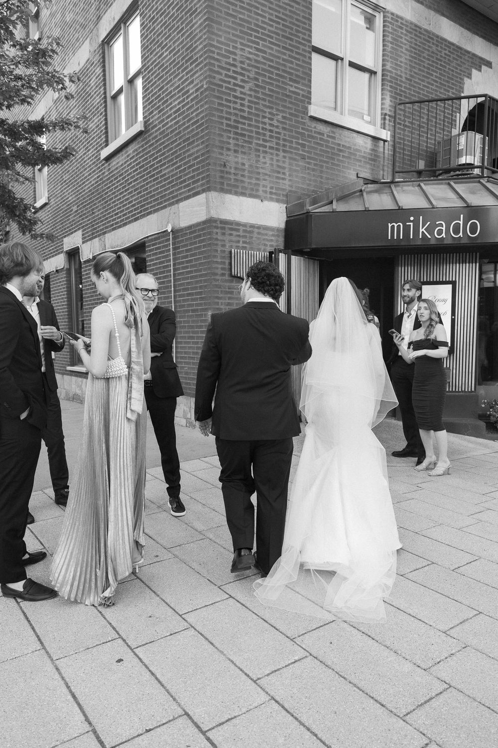 Black and white photo of a bride and groom walking toward a venue entrance with wedding guests outside.
