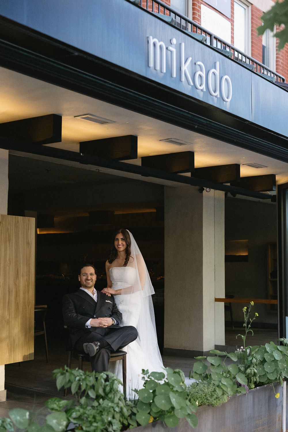 Bride and groom posing outside Mikado restaurant, groom seated in suit, bride standing in white gown and veil.