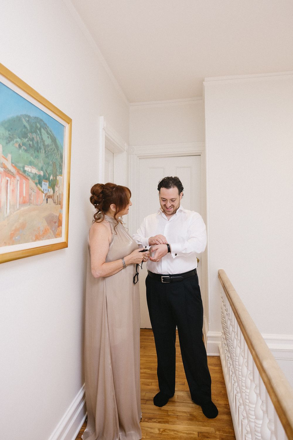 Couple getting ready in a hallway, woman in taupe gown helping man with cufflinks near a colorful framed painting.